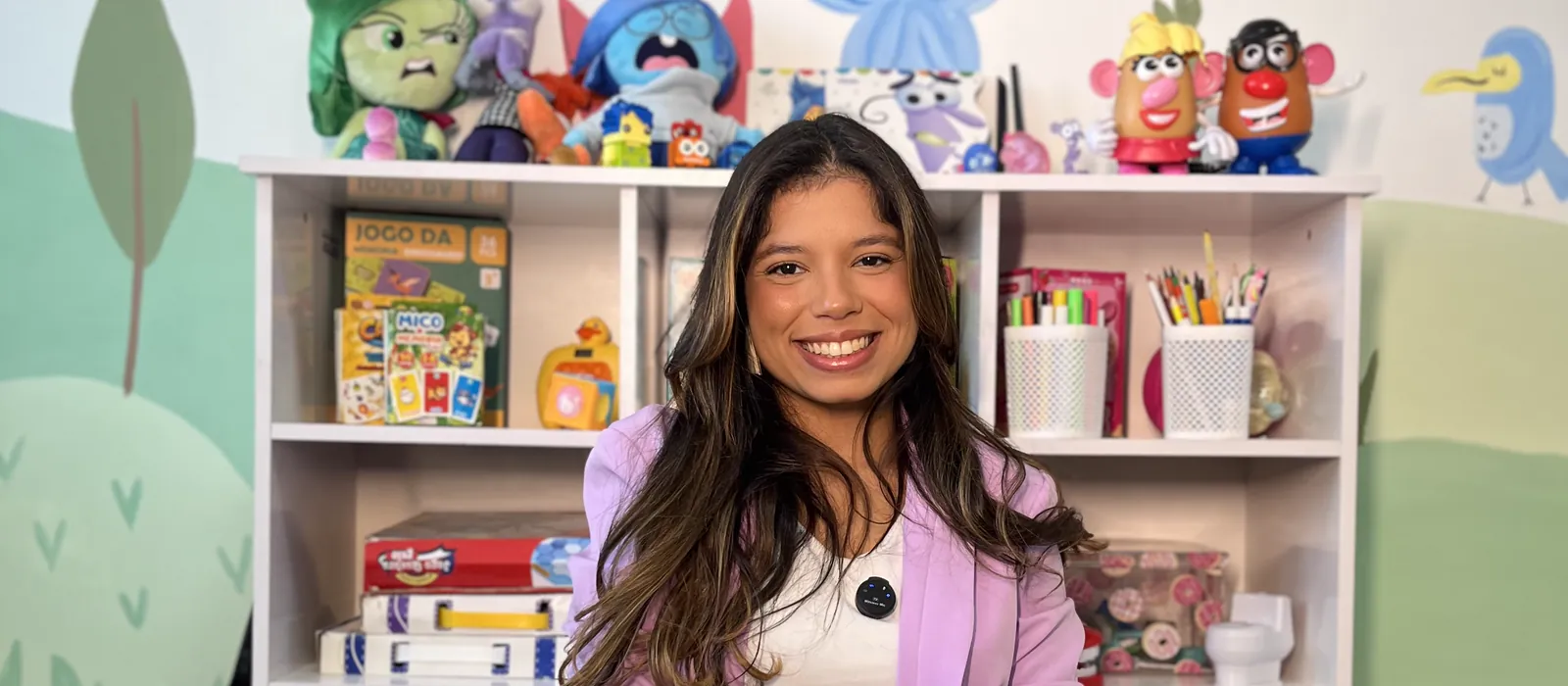 Jessica Costa sorrindo no consultório do Vertice Mall, Recreio dos Bandeirantes — estante com pelúcias dos personagens Divertida Mente, jogos pedagógicos e mural infantil ao fundo